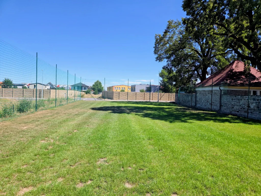Zelená plocha, čerstvě zasetá, na horizontu modré nebe, u zelené plochy je zeď