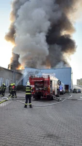 Hasiči zasahují u průmyslové budovy v Čelákovicích. Ze střechy stoupá hustý šedý kouř.