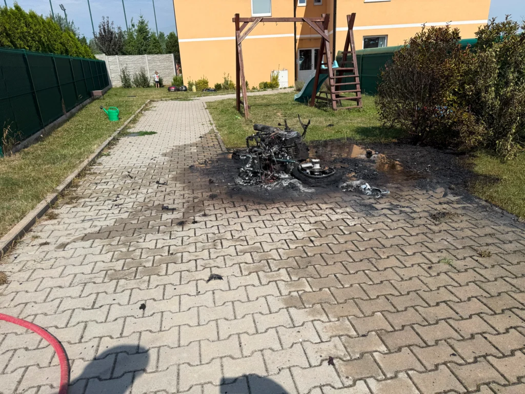 Na vydlážděném dvoře před žlutou budovou leží ohořelý motocykl