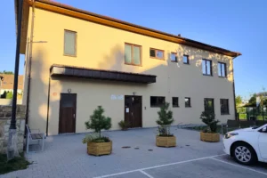 Budova dětské skupiny. Dvoupatrová, žluté barvy, před budovou okrasné keře a parkoviště.