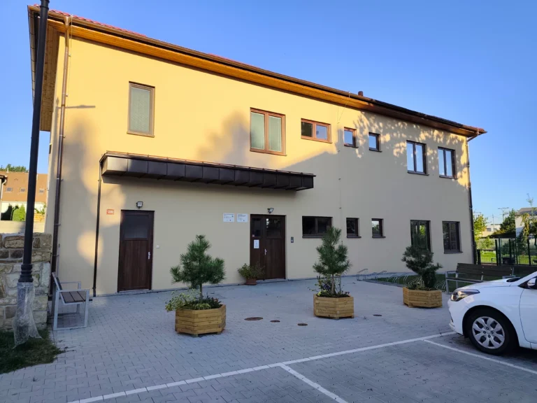 Budova dětské skupiny. Dvoupatrová, žluté barvy, před budovou okrasné keře a parkoviště.