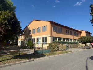 Budova mateřské školy, oranžová, modré nebe, zelený plot a kousek zahrady.