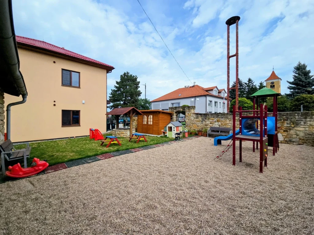 Zahrada u budovy s béžovou fasádou, jsou tam dětské prvky