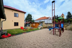 Zahrada u budovy s béžovou fasádou, jsou tam dětské prvky